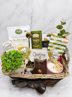 A wicker gift basket filled with assorted gourmet snacks, tea, crackers, a candle, and small greenery accents, photographed on a light marble surface against a white marble background.