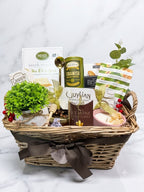 A wicker gift basket filled with assorted gourmet snacks, chocolates, and a candle, decorated with a brown ribbon and small greenery, set against a white marble background.